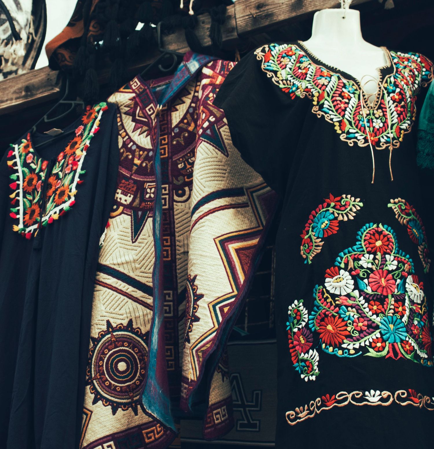 Black traditional dresses with colorful floral embroidery and geometric patterned garments hang on display in a shop.