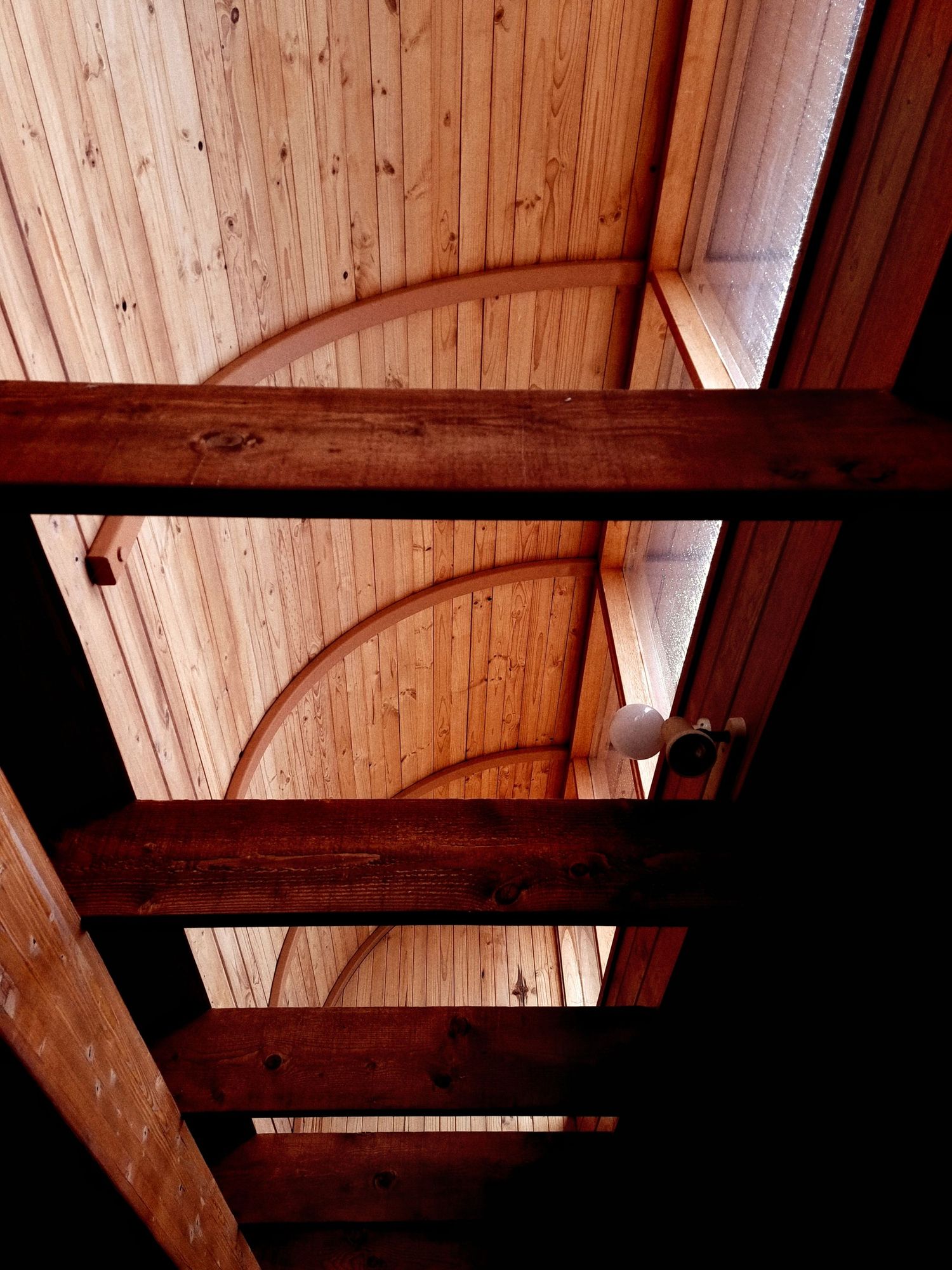 Warm wooden ceiling with arched beams and exposed dark timber rafters, lit by a skylight and a small hanging light.