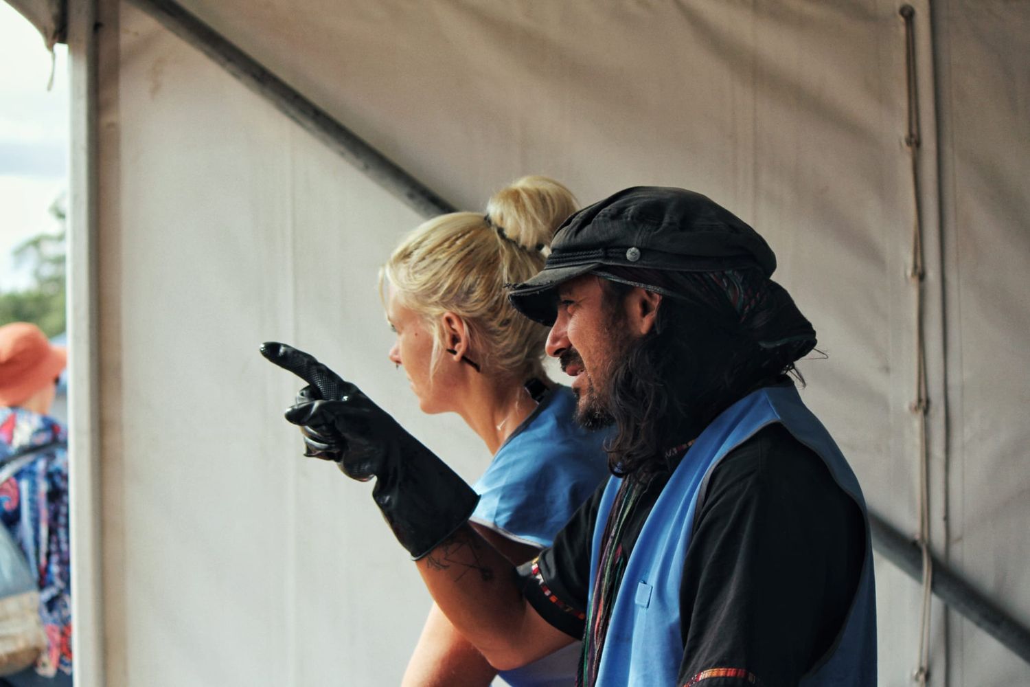 Man in a flat cap and blue vest points ahead while a blonde woman beside him looks on under a white canvas tent.