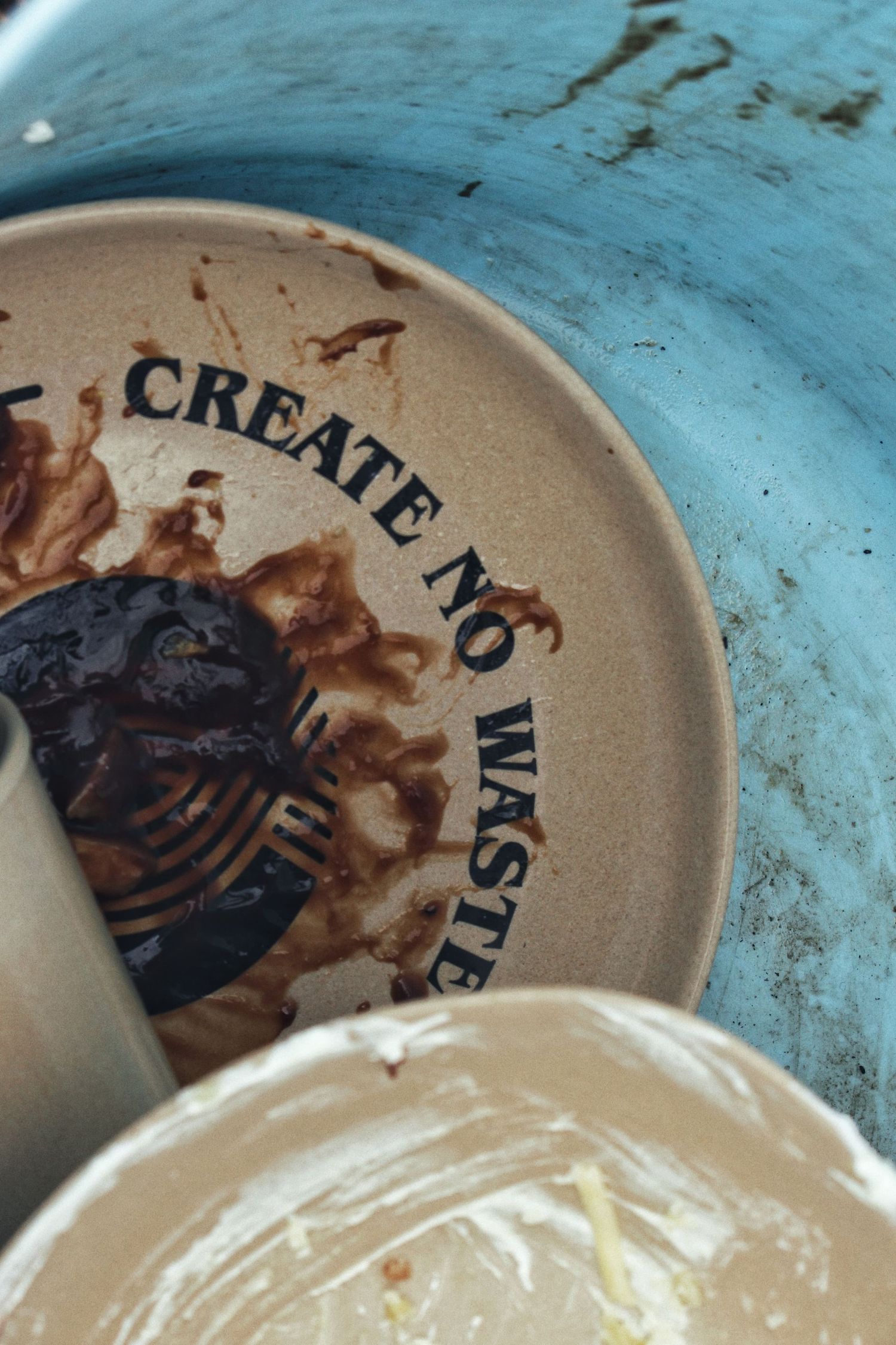 A dirty ceramic plate stamped with the words Create No Waste sits in a blue washing tub, with food scraps and sauce smears.