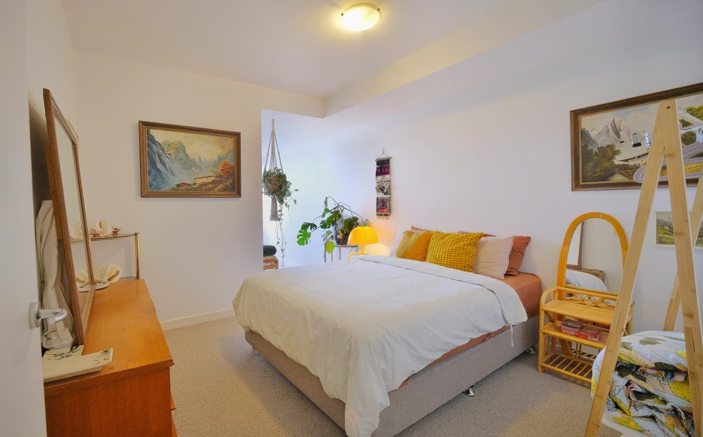 Bright, cozy bedroom with a white-bedded double bed, yellow cushions, wooden dresser, framed landscape artwork and indoor plants.