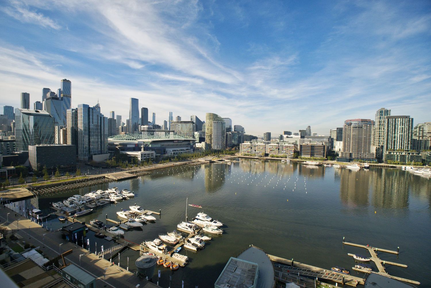 Waterfront city skyline with high-rise buildings overlooking a marina filled with moored boats under a blue sky with wispy clouds.