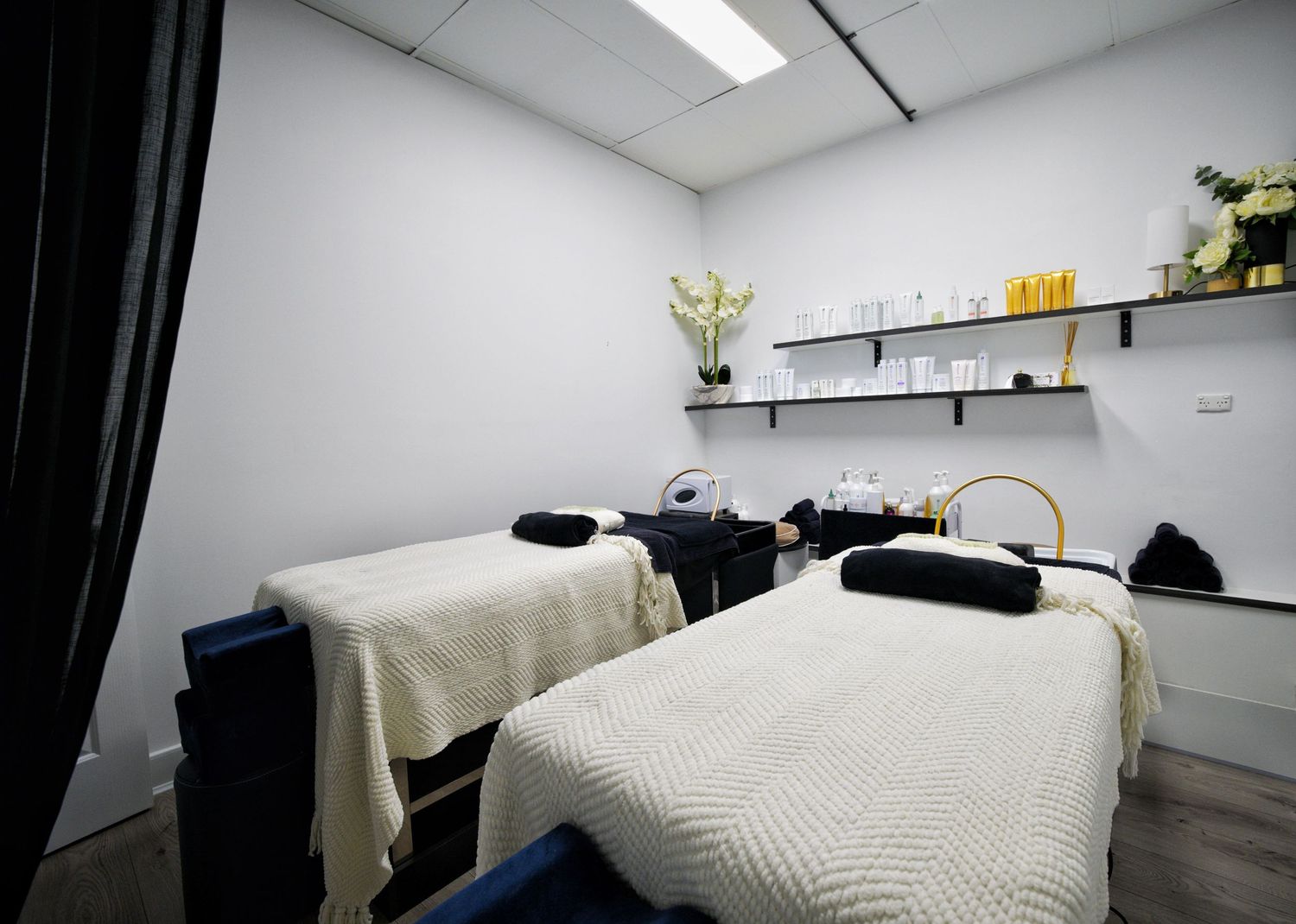 Bright treatment room with two massage beds with cream blankets and shelves of skincare products on the wall.