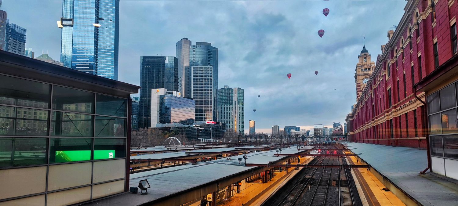 Hot air balloons float over a city skyline at dusk as a train platform and brick buildings line the railway tracks below.