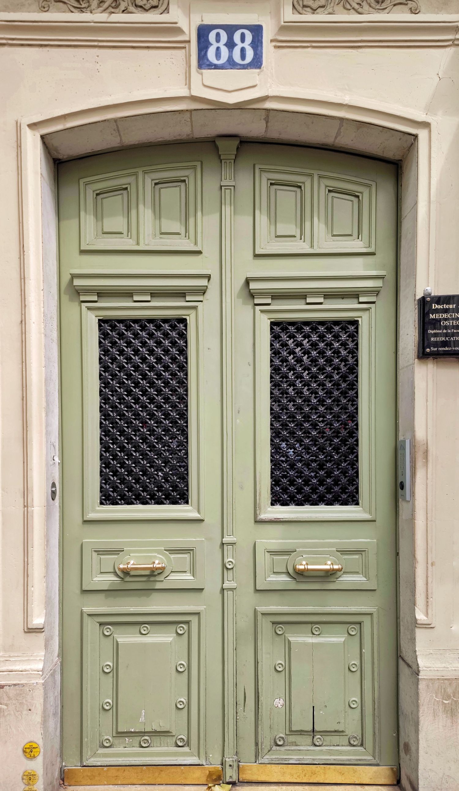 Ornate sage green double door with black lattice windows and brass handles beneath a blue number 88 plaque on a cream Parisian facade.