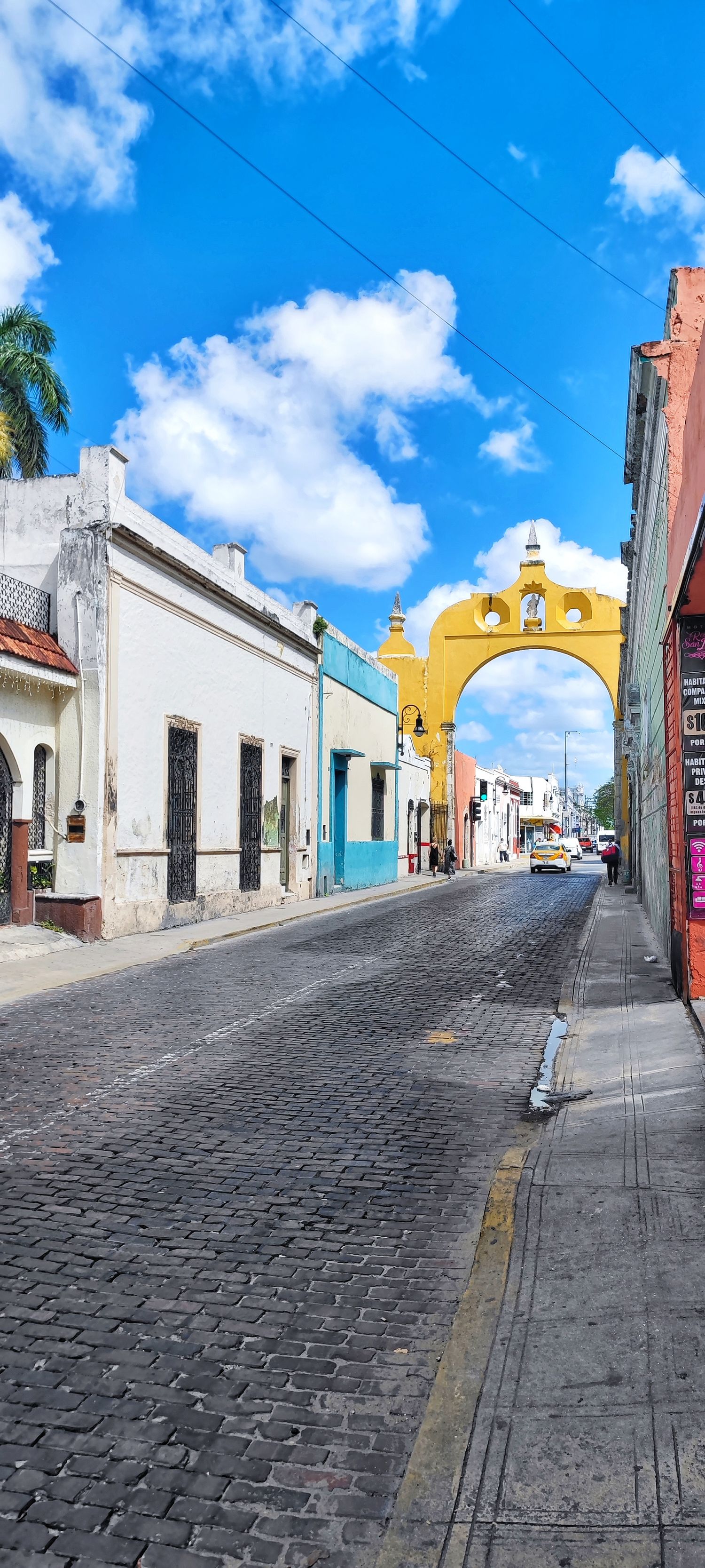 Cobblestone street lined with colorful colonial buildings leads to a golden arch beneath a bright blue sky with clouds.