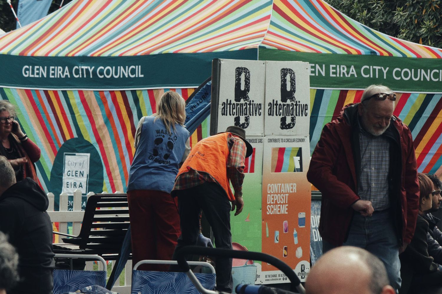 Event staff and visitors stand beside a colourful striped Glen Eira City Council tent displaying container deposit scheme signs.