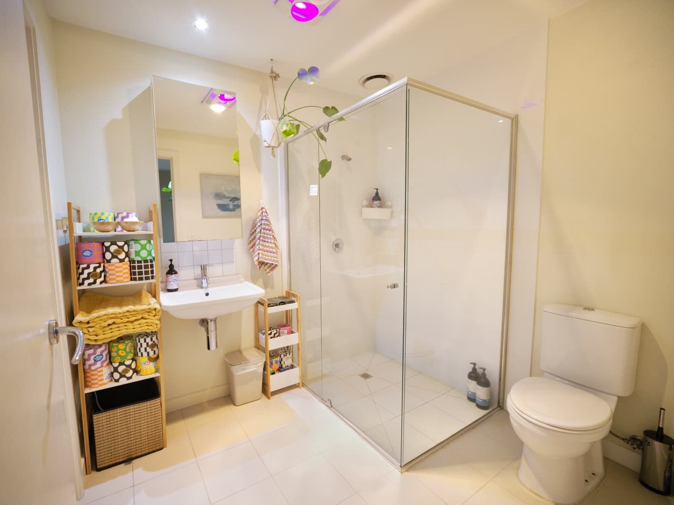 Modern bathroom with a glass shower enclosure, wall-mounted sink, wooden shelving with colorful baskets, toilet and hanging plant.
