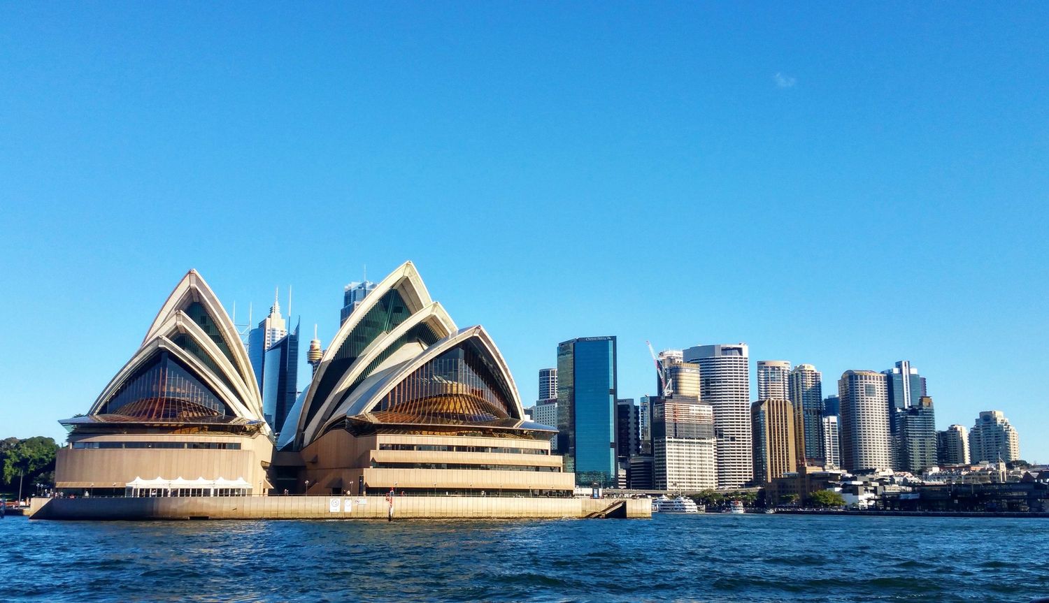 The Sydney Opera House and the city skyline stand along the harbor under a clear blue sky, seen from the sparkling water.