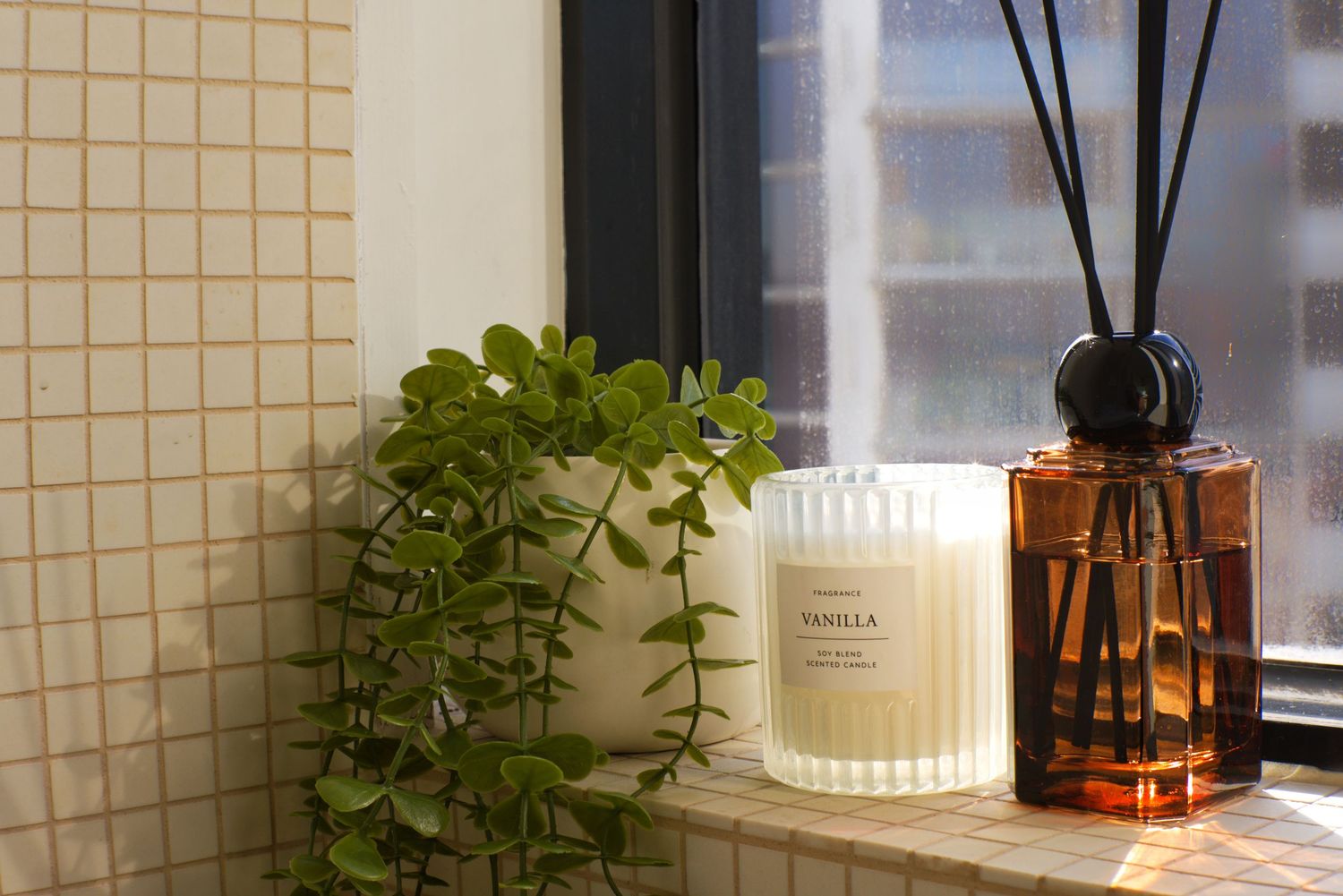 Windowsill with a green potted plant, a vanilla scented candle and a reed diffuser beside beige mosaic wall tiles.
