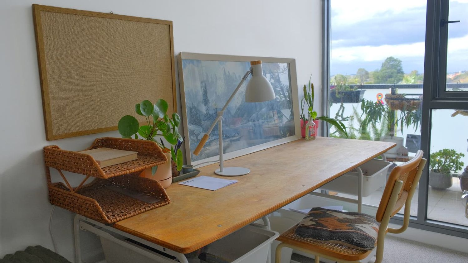 Timber work desk with a lamp, woven storage trays and a plant beside a window with a balcony and city view.