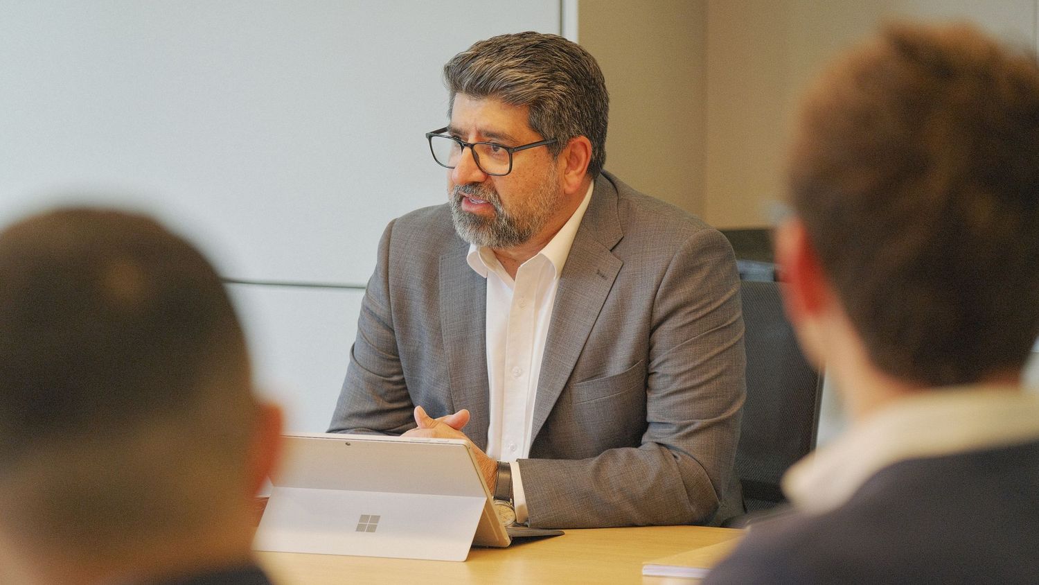 A bearded man in glasses and a gray blazer speaks during a business meeting while using a tablet at a conference table.