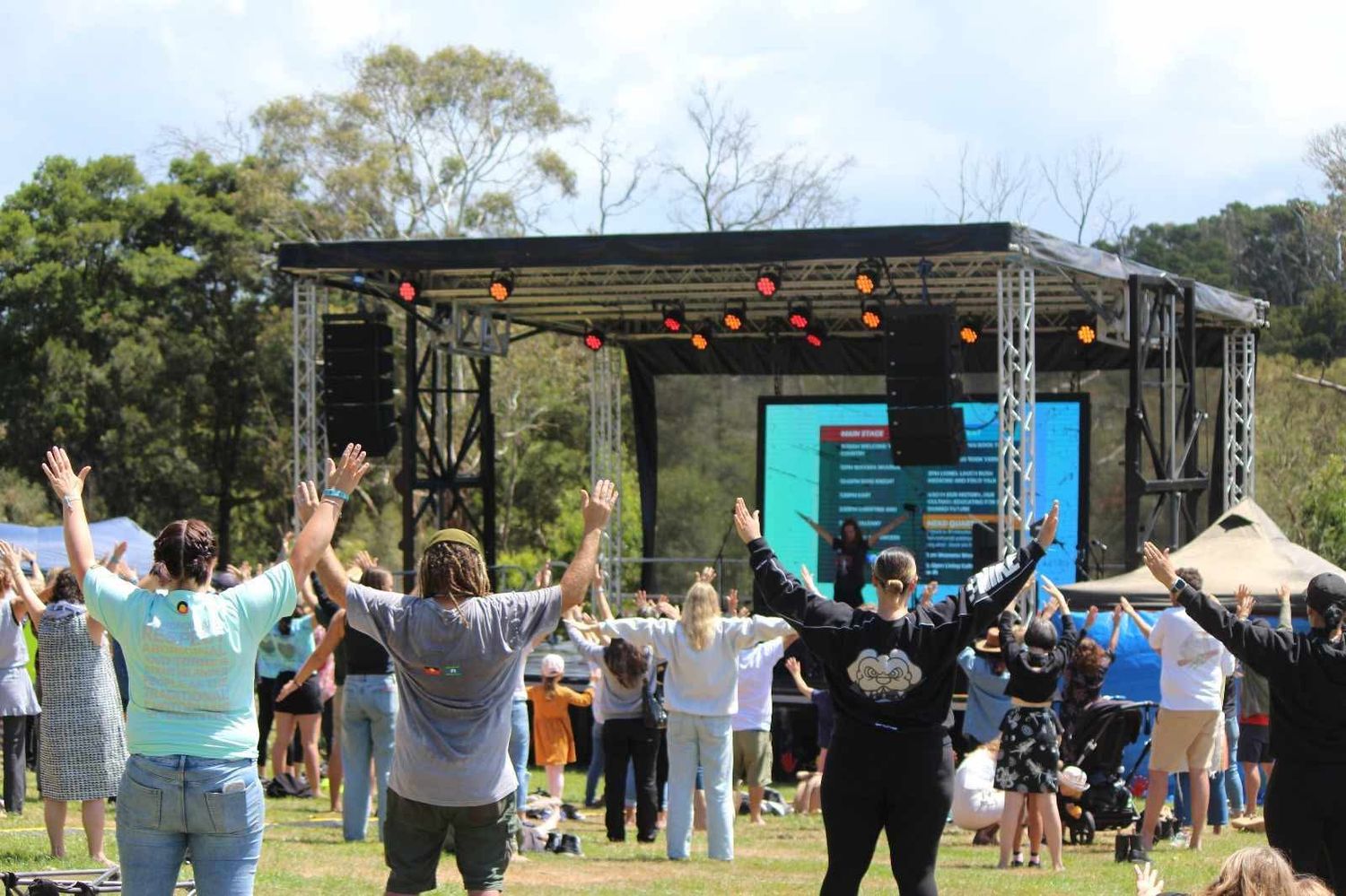A crowd with raised arms worships on a grassy field in front of an outdoor stage with lights and a large screen surrounded by trees.