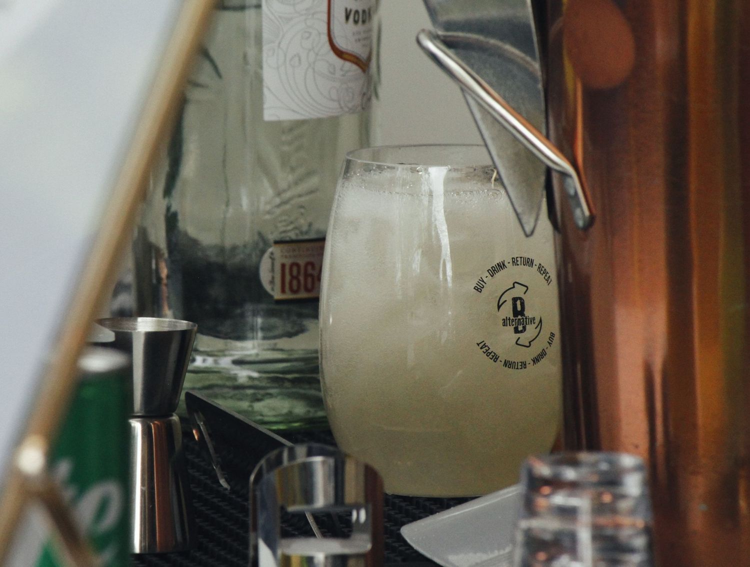 A frosty glass of cocktail sits on a bar counter beside a metal jigger and liquor bottles.