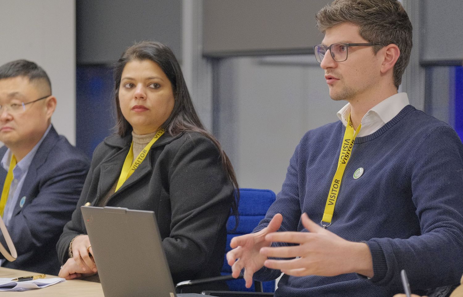 A man in glasses and a lanyard gestures while speaking to colleagues at a conference table with a laptop during a workshop.
