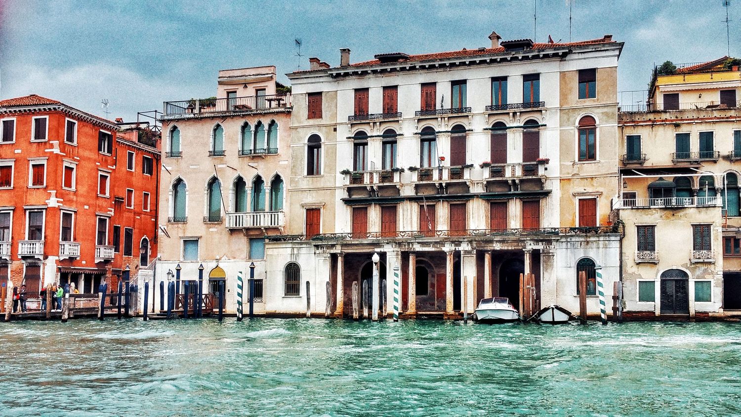 Historic palazzo facades in orange, cream and red line the Grand Canal in Venice, with rippling green water in the foreground.