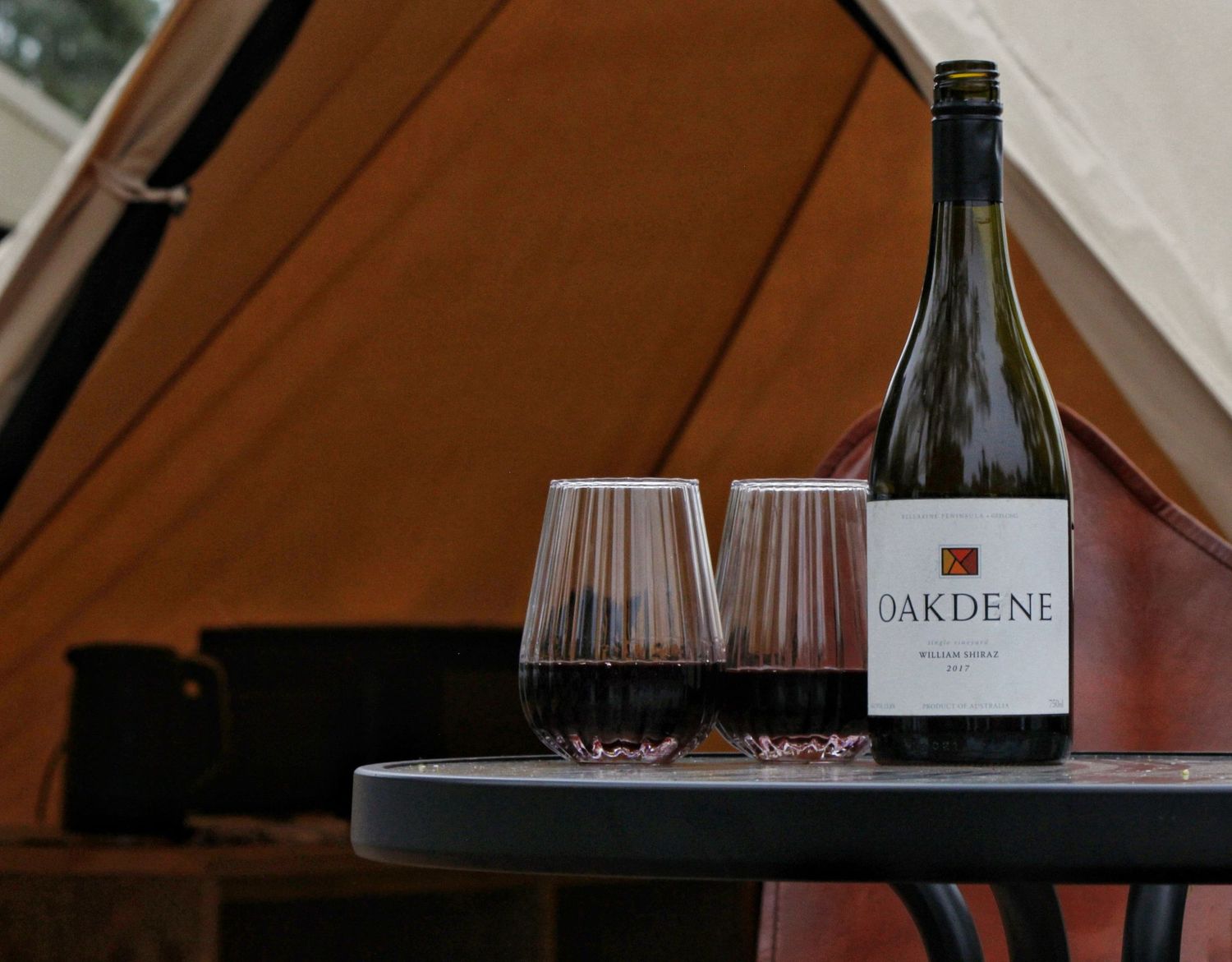 A bottle of Oakdene wine and two filled glasses sit on a small round table in front of a canvas bell tent.