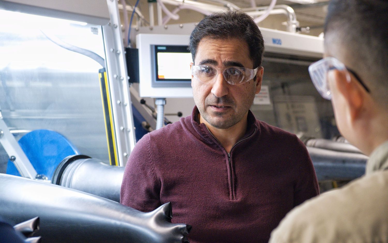 A man in glasses and a maroon sweater listens attentively beside a laboratory glovebox with ducting overhead.