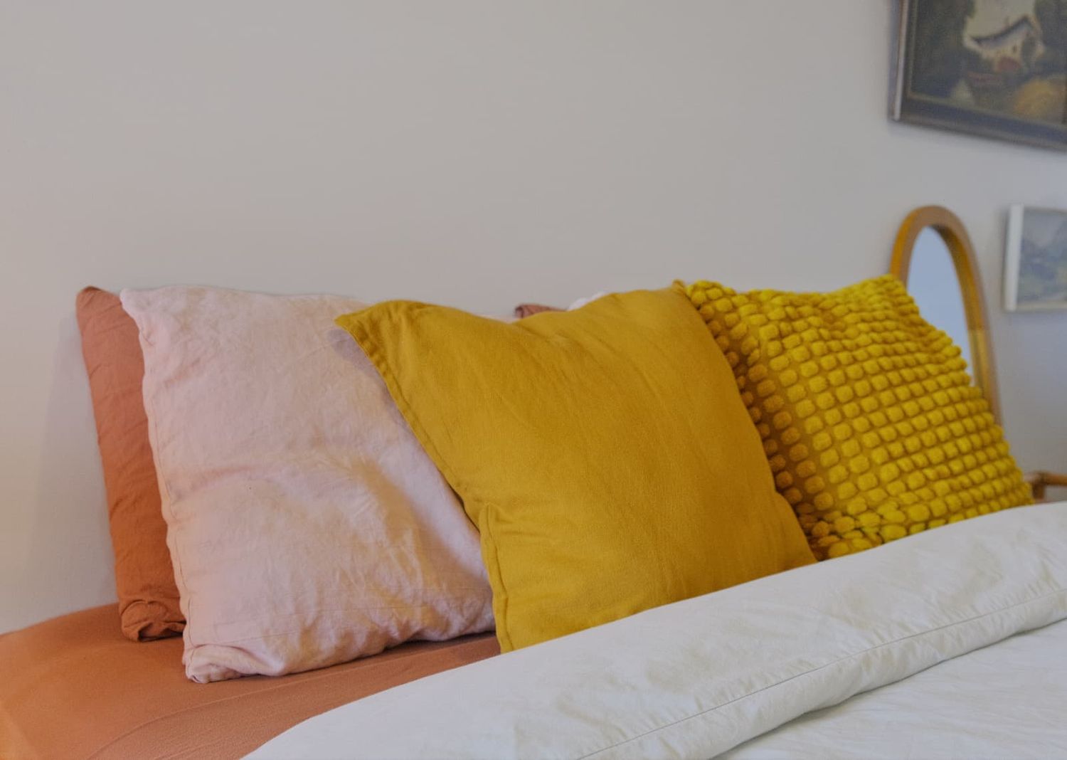 Close-up of mustard yellow, blush pink and textured cushions piled on a bed with white linen and a cane headboard.
