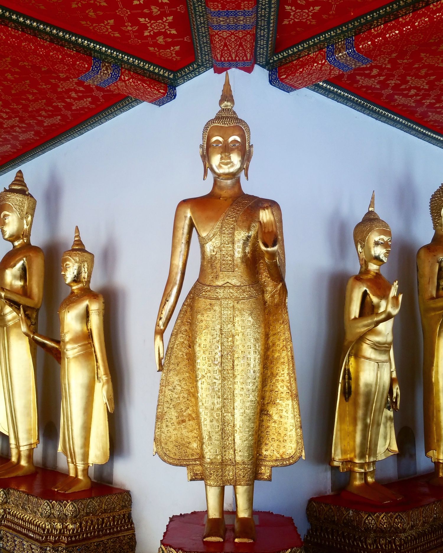Golden standing Buddha statues in a temple hall with a red ornate ceiling, with the central figure framed by smaller gilded Buddhas.