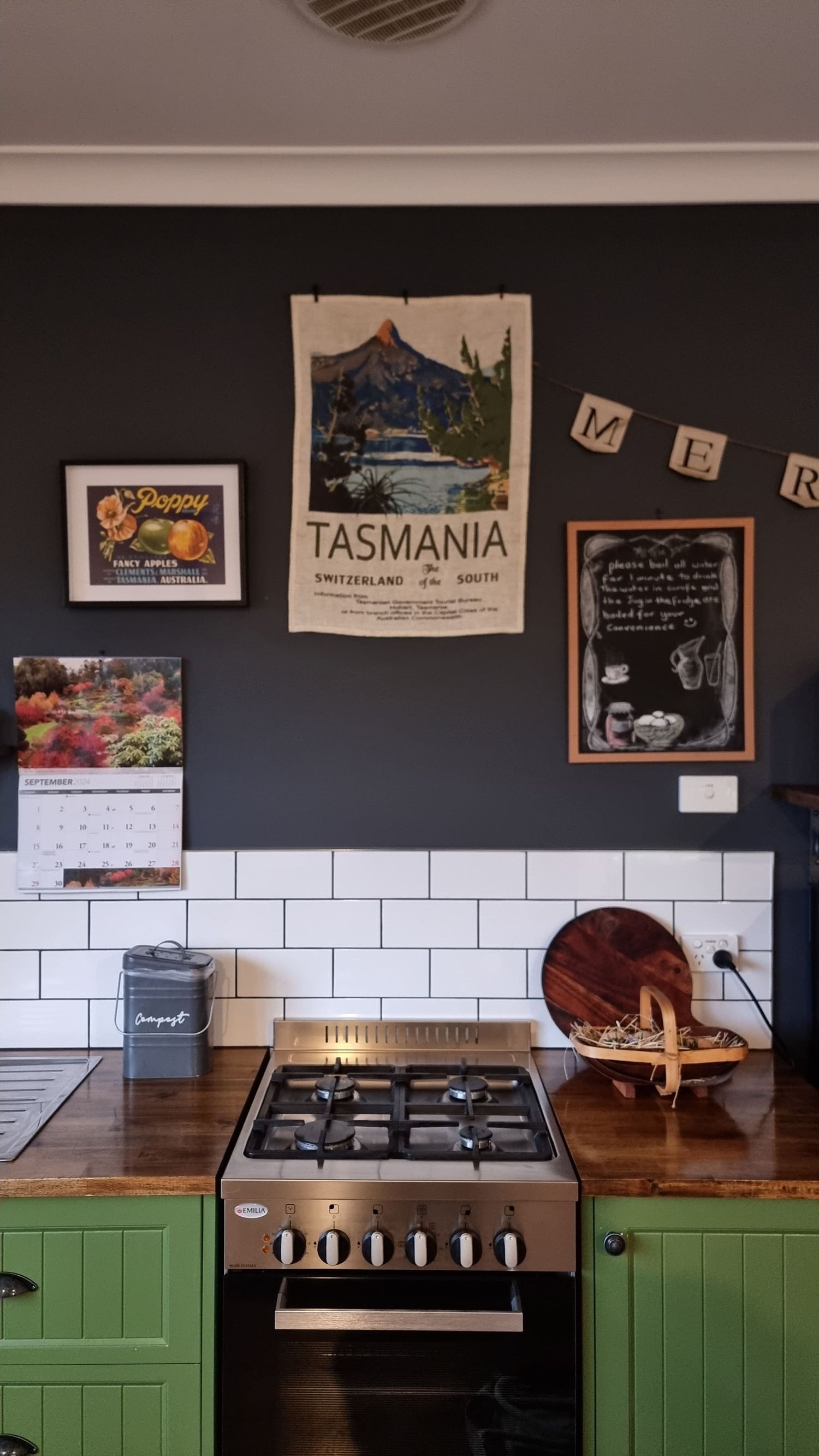 Cozy farmhouse kitchen with green cabinets, a gas stove, wooden benchtops, white tiles and a vintage Tasmania poster on a dark wall.