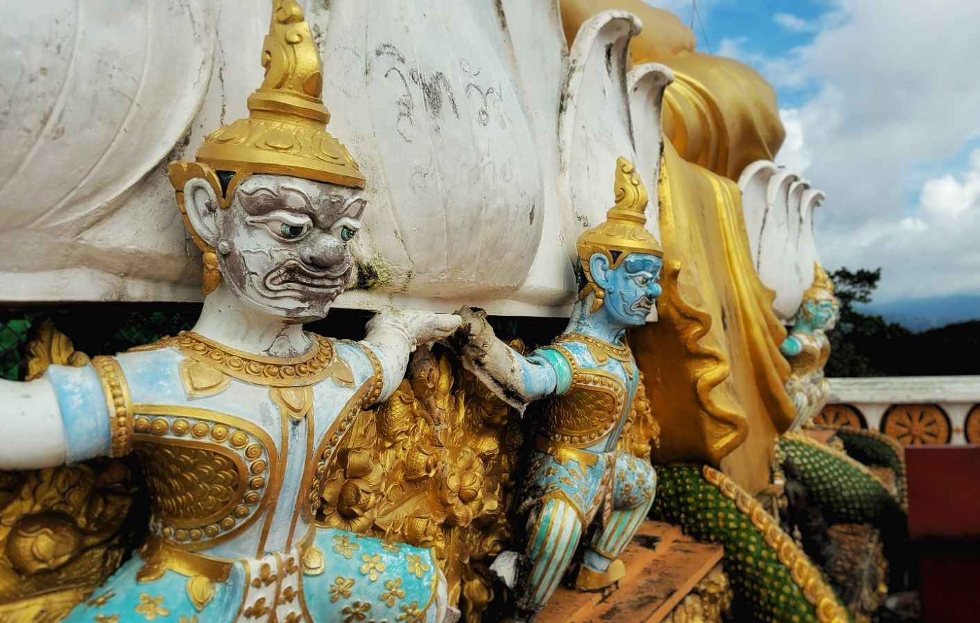 Colorful temple statues of mythical guardian figures in gold and turquoise costumes beside a large white Buddha figure.