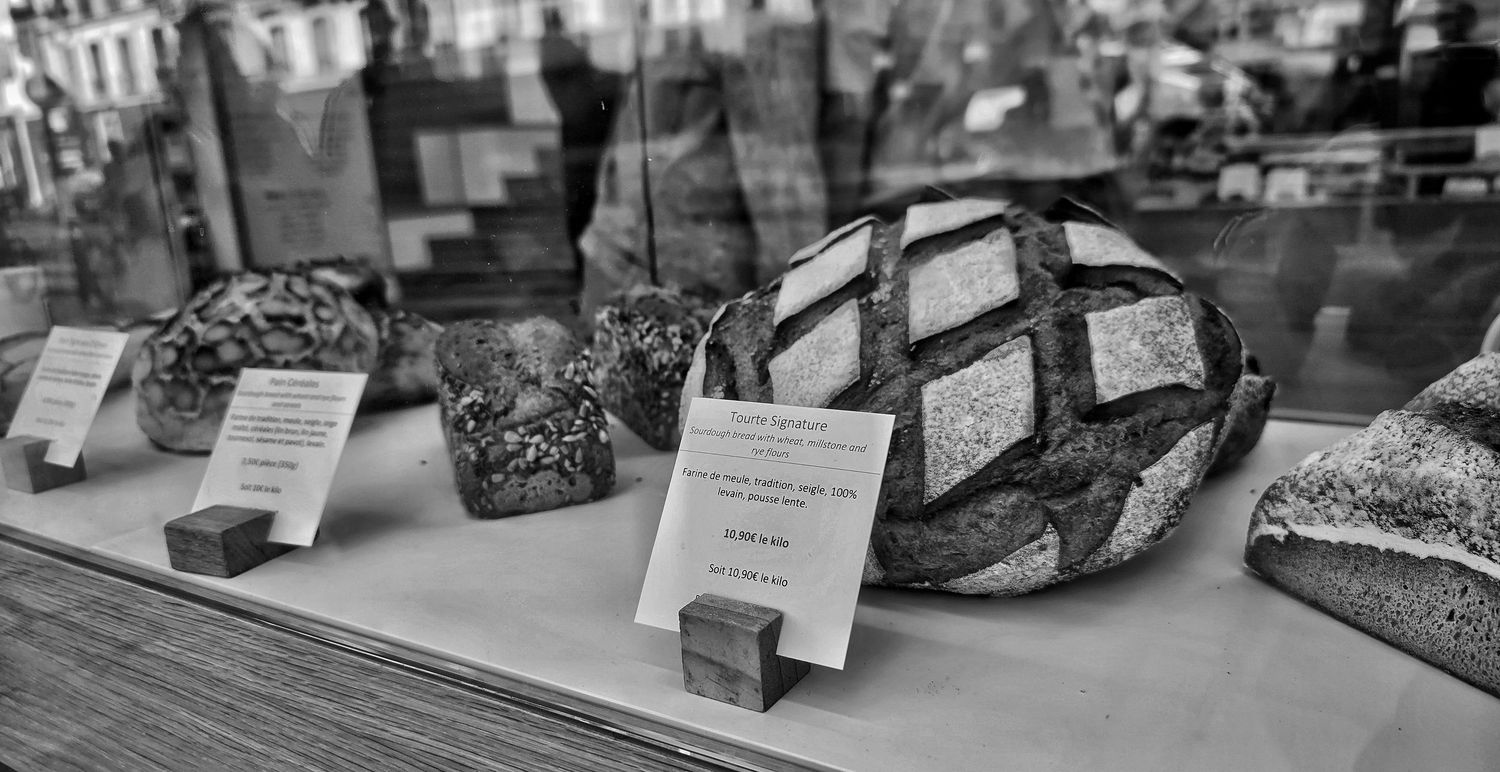 Black and white display of artisan sourdough loaves with scored crusts and small price cards behind a bakery shop window.