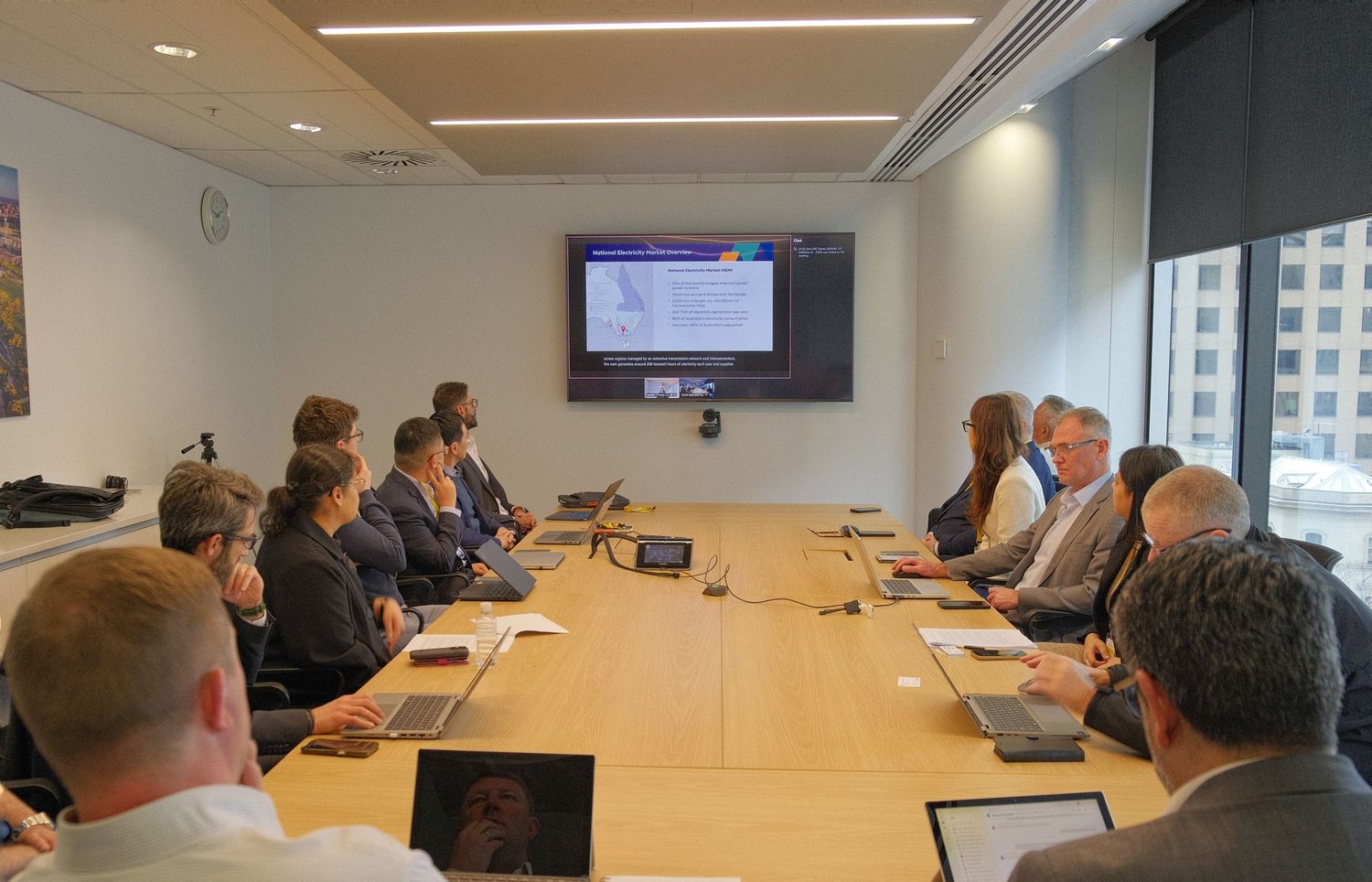 Professionals sit around a long wooden conference table with laptops while a presentation is displayed on a wall-mounted screen.
