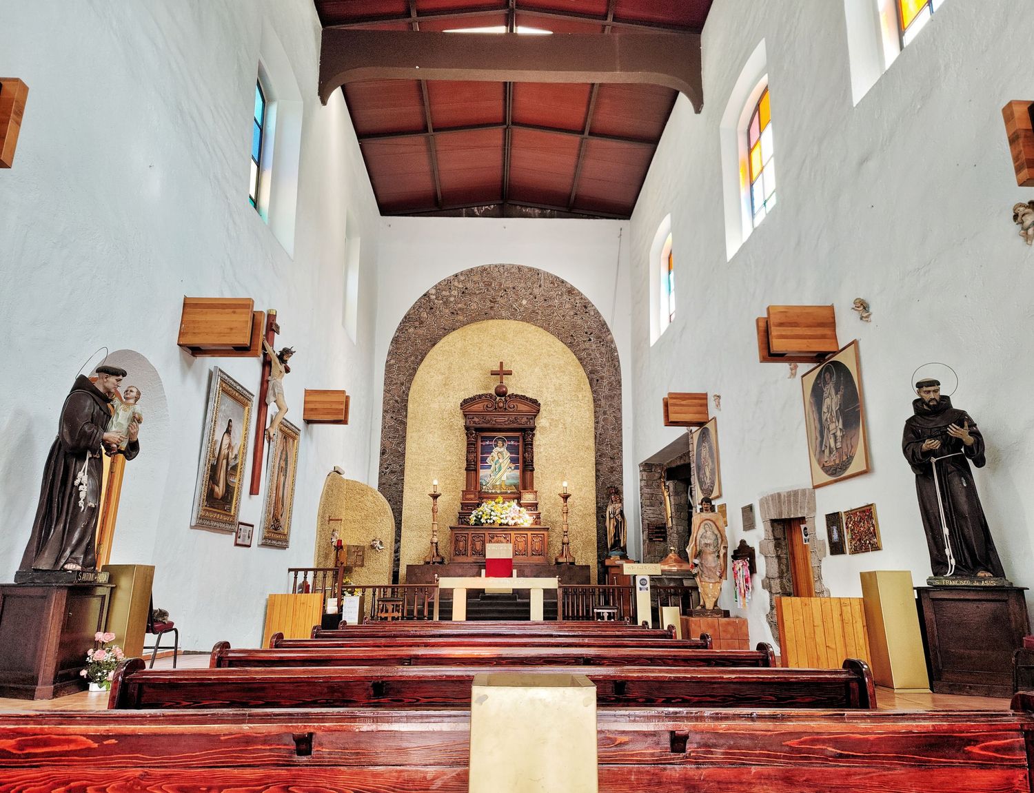 Quiet chapel interior with wooden pews, a domed stone arch behind the altar, religious statues and stained glass windows.