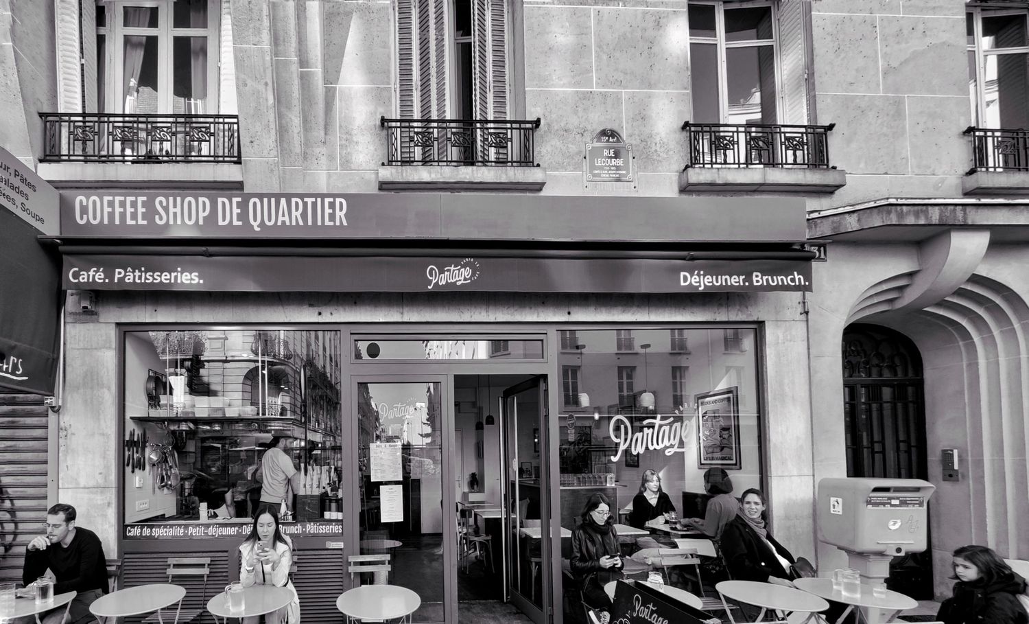 Black and white view of a Parisian coffee shop storefront with patrons seated at café tables inside and outside the window.