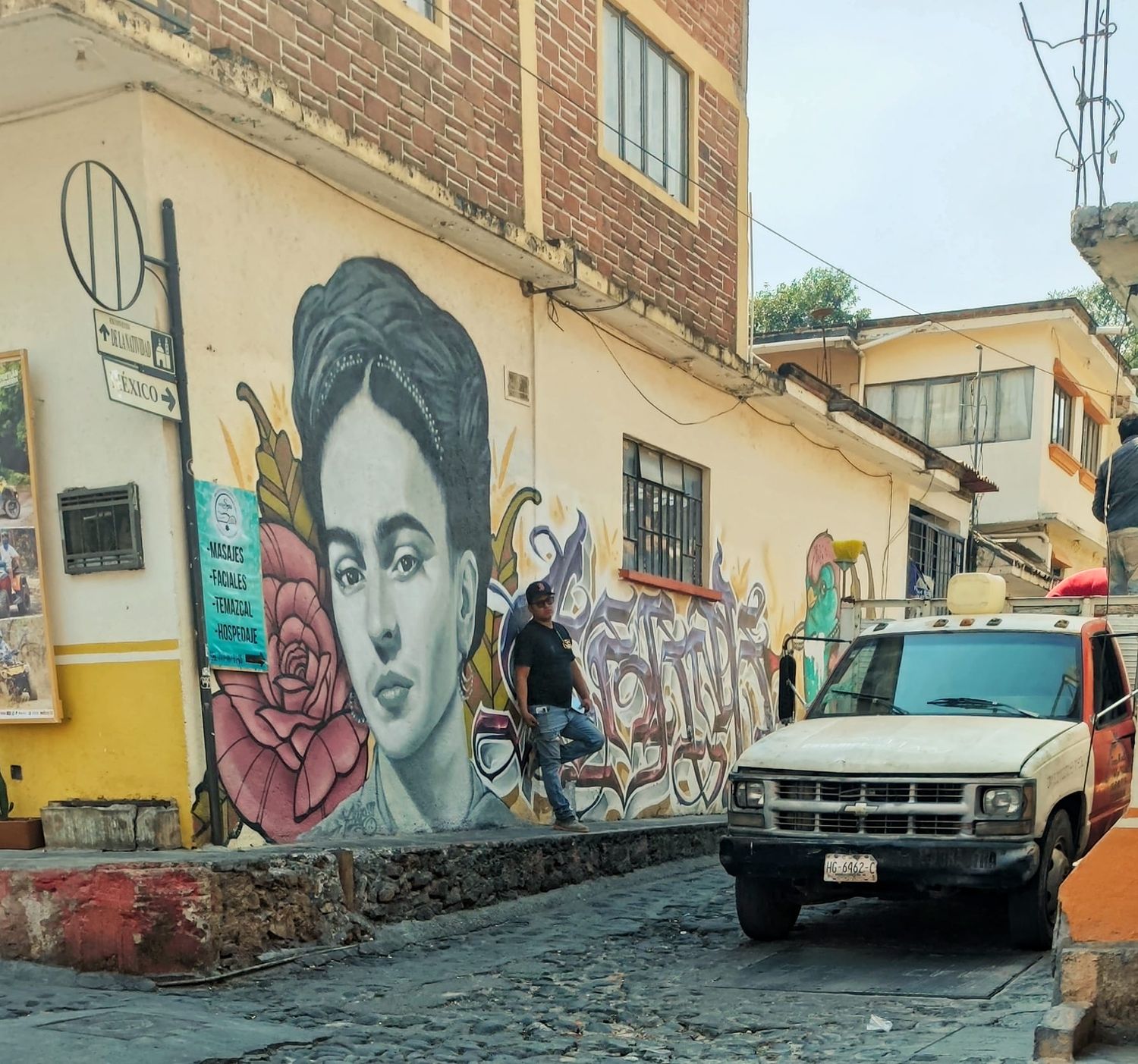 A large mural of a woman with a rose adorns a yellow building wall beside a cobblestone street where a white pickup truck is parked.