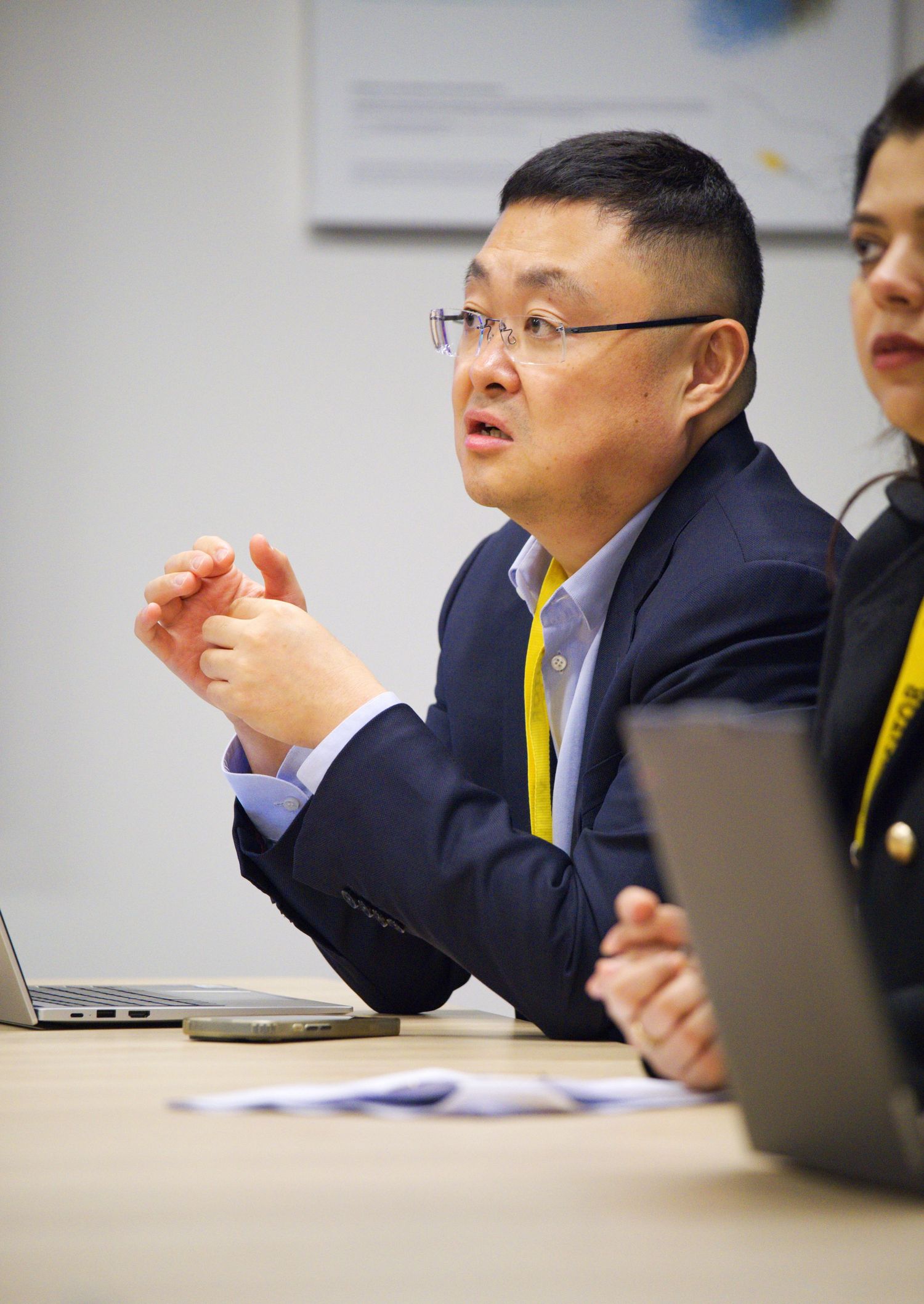 A man in glasses and a navy blazer gestures while speaking at a conference table with laptops and papers.