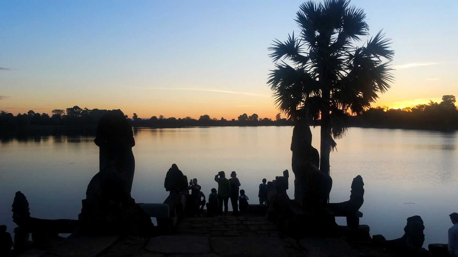 Silhouetted visitors and stone statues beside a calm lake at sunset with a palm tree and glowing orange sky in Angkor, Cambodia.