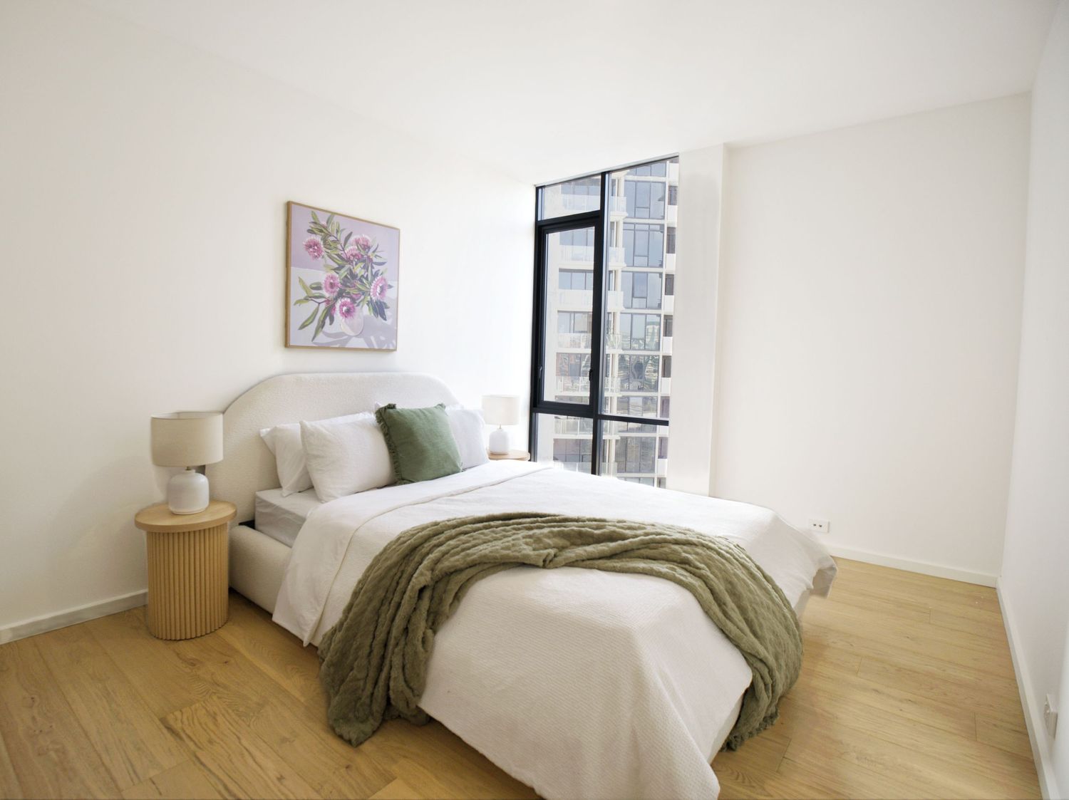 Light-filled bedroom with a double bed, olive green throw, bedside table, floral artwork and a floor-to-ceiling window.