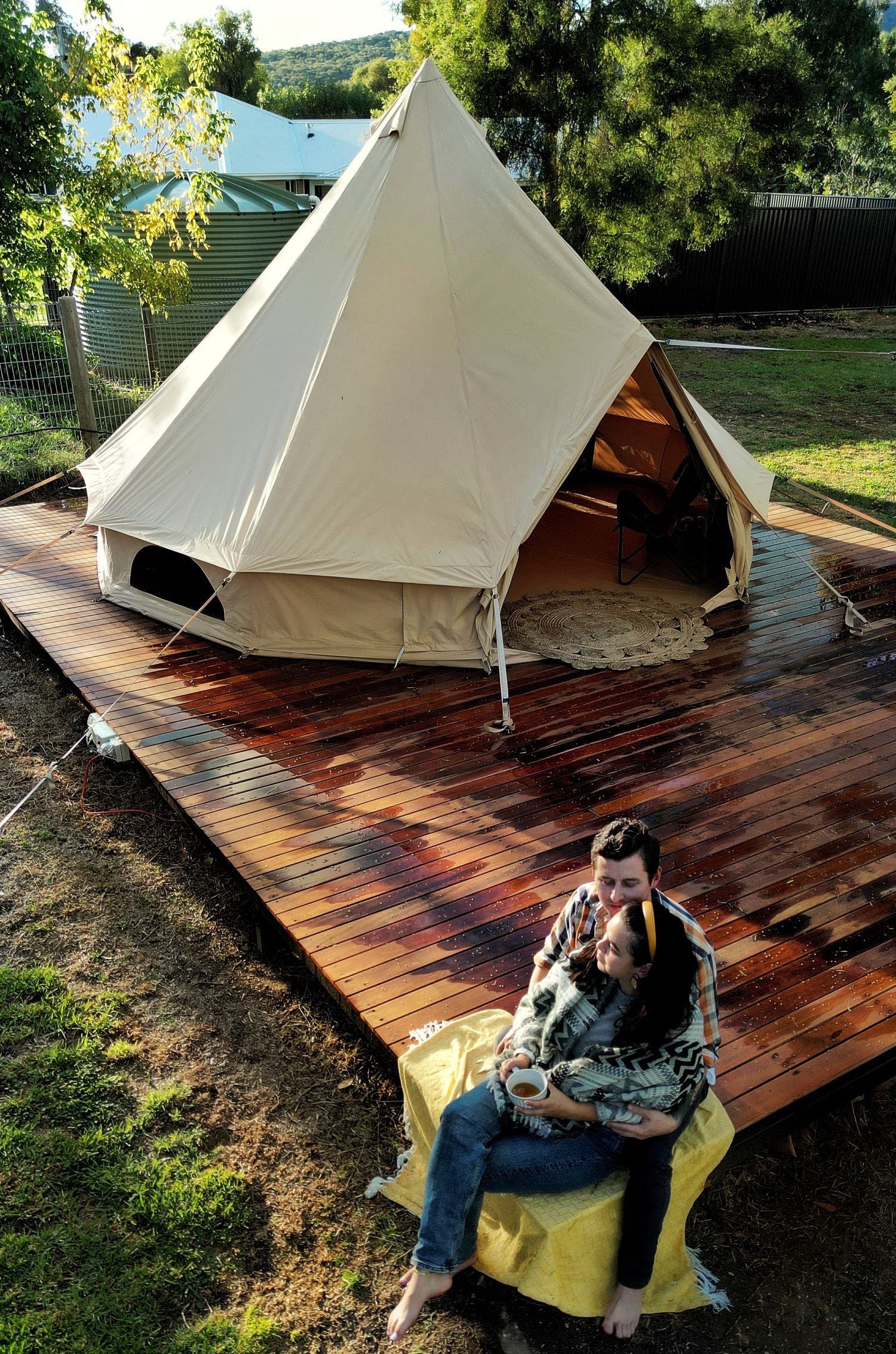 A couple cuddles on a yellow blanket on a wet timber deck in front of a cream canvas bell tent surrounded by trees.