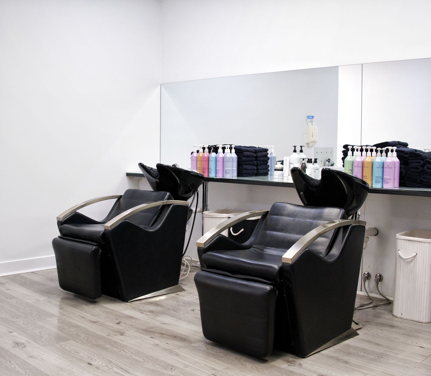 Two black reclining shampoo chairs with basins in front of a mirrored wall lined with colorful salon shampoo and conditioner bottles.