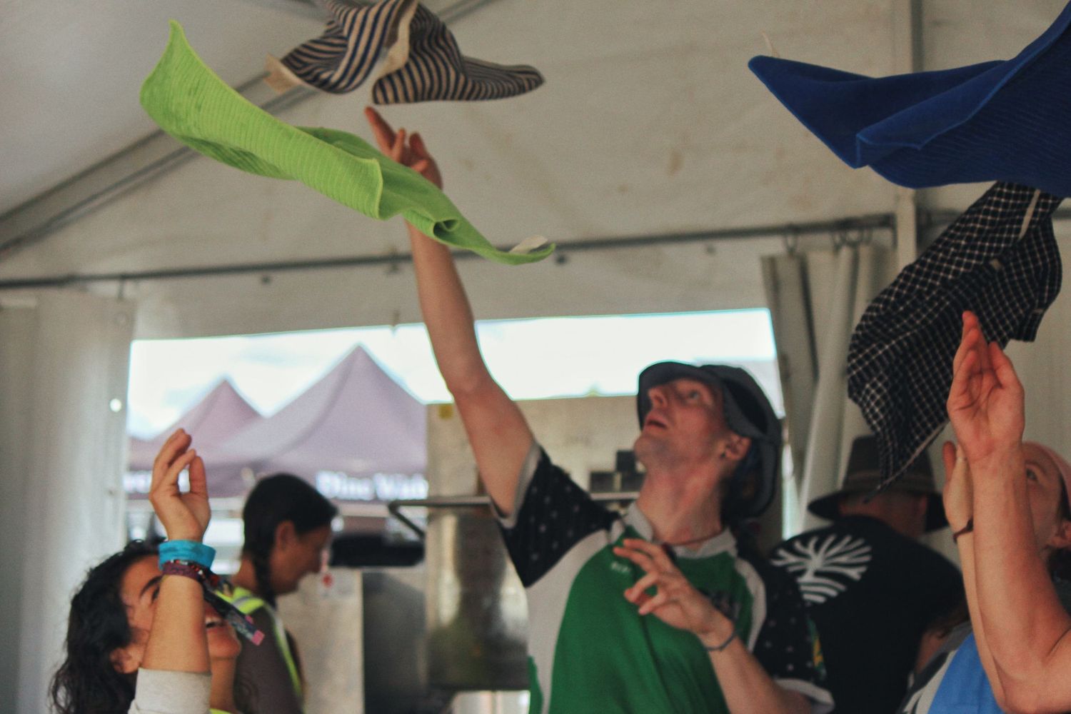 Festival goers under a tent toss colorful folded paper hats into the air, laughing and reaching up together.