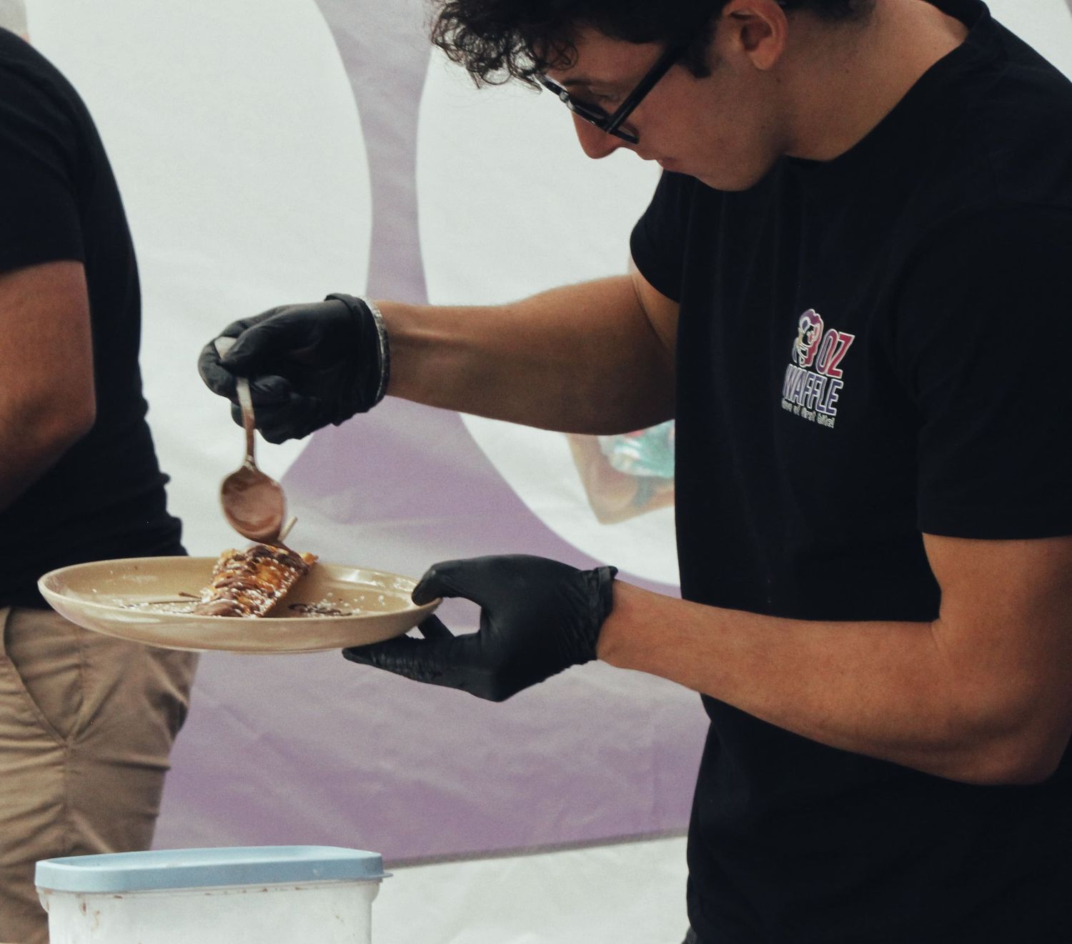 A food vendor in black gloves and a black t-shirt spoons sauce onto a plate of grilled food at an outdoor event.