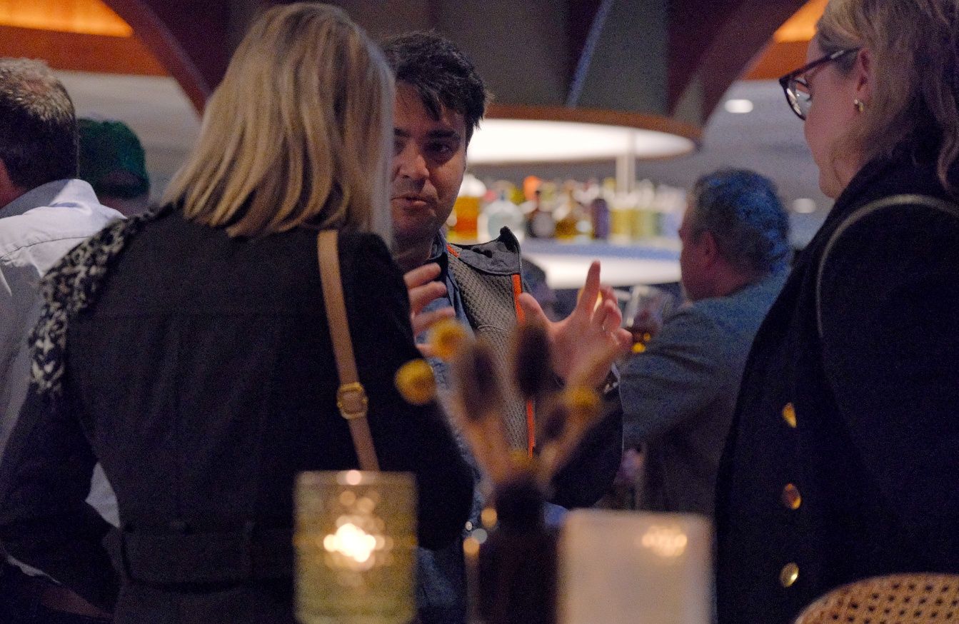Guests chat and gesture during a networking event in a warmly lit bar, with a candle and dried flowers in the foreground.