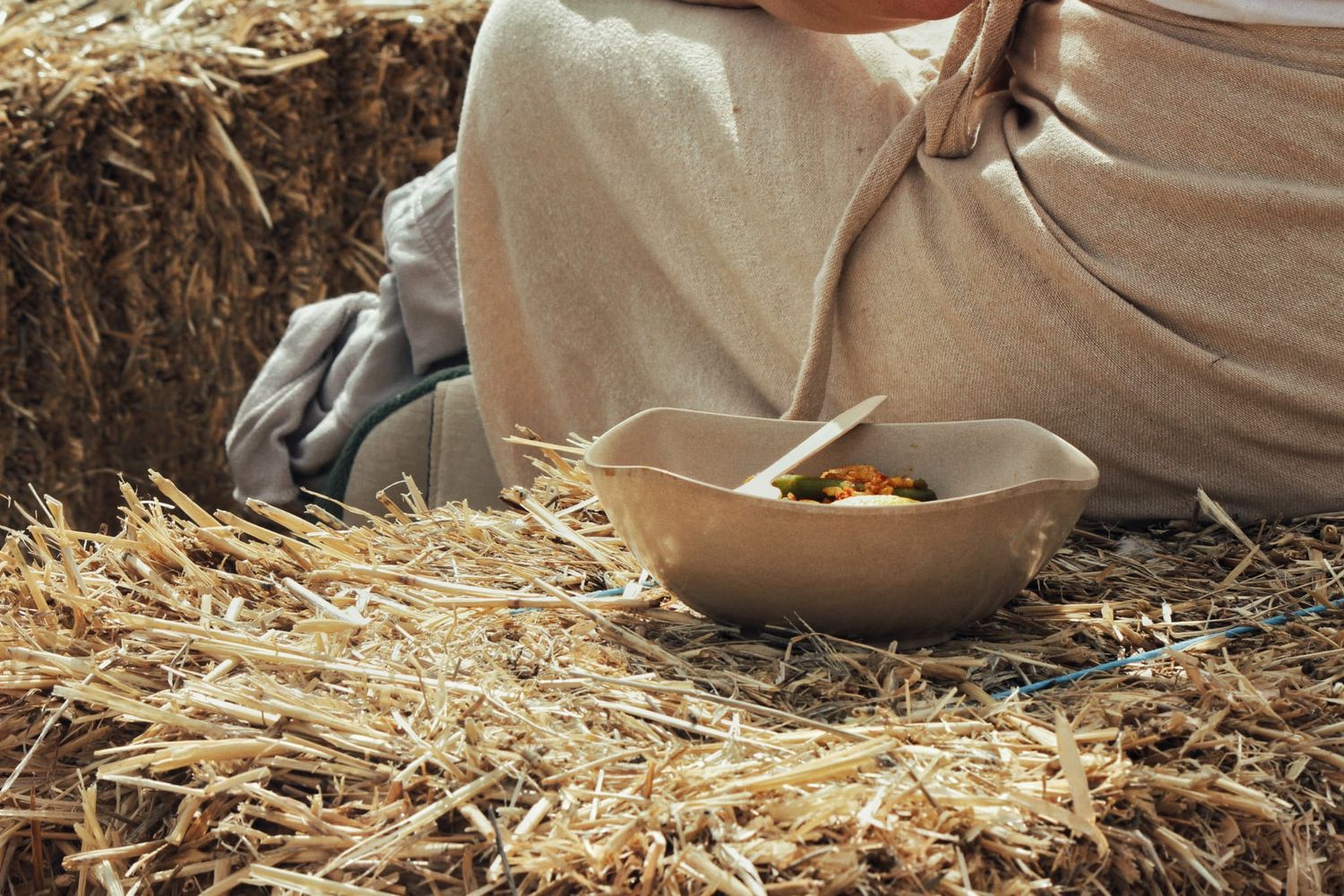 Person sitting on straw bales rests a compostable bowl of food on the straw ground at an outdoor event.