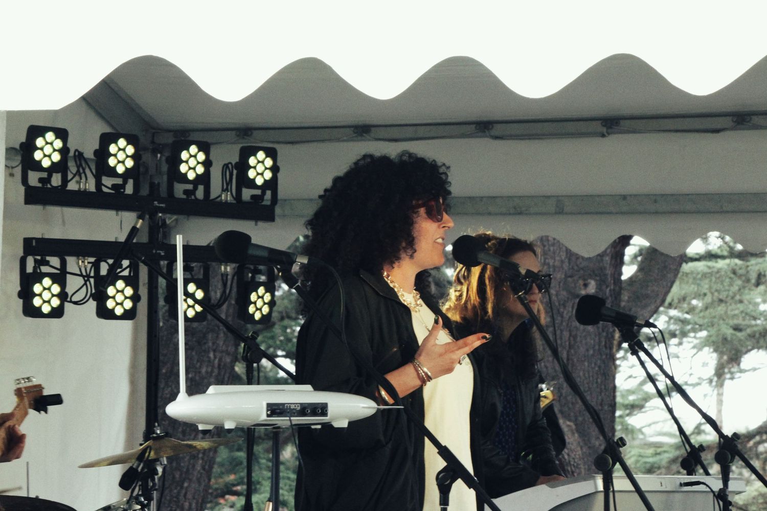 A curly-haired singer performs at a microphone on a covered outdoor stage lit by rows of bright stage lights.