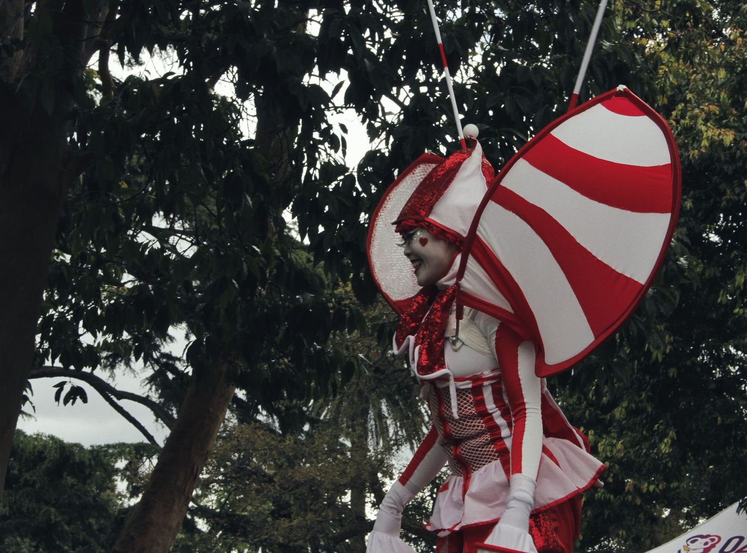 A performer in a red and white striped costume with large wing-like panels stands among trees at an outdoor festival.