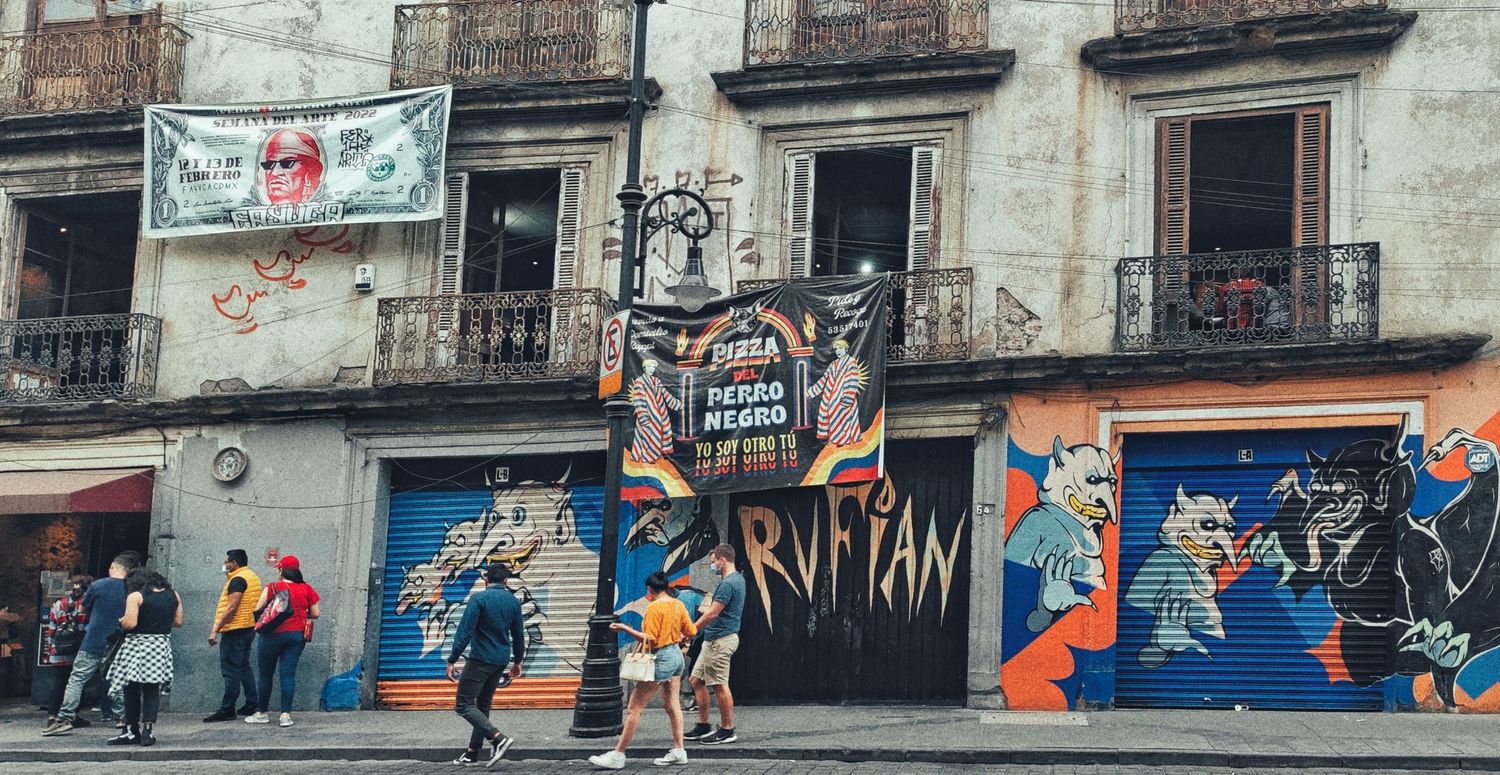 Pedestrians walk past a colorful street art mural and wrought iron balconies on a weathered building in a lively city street.