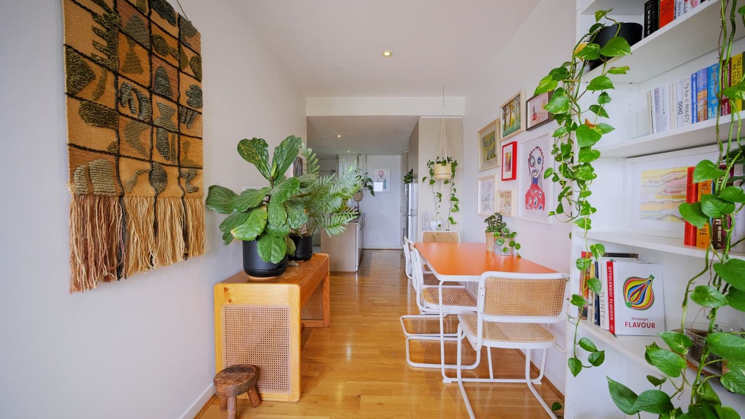 Bright apartment hallway with a woven wall hanging, leafy houseplants, a bookshelf and an orange dining table with white chairs.