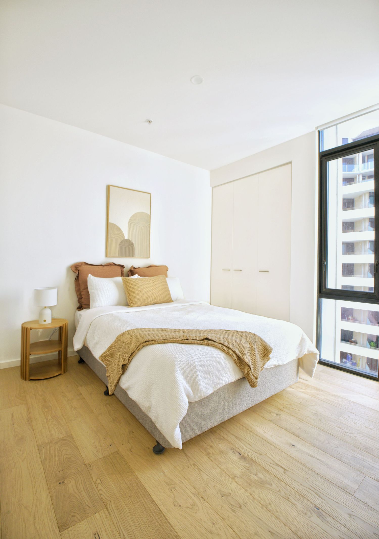 Light-filled bedroom with a double bed, tan and rust bedding, timber floors, built-in wardrobe and a side table lamp.