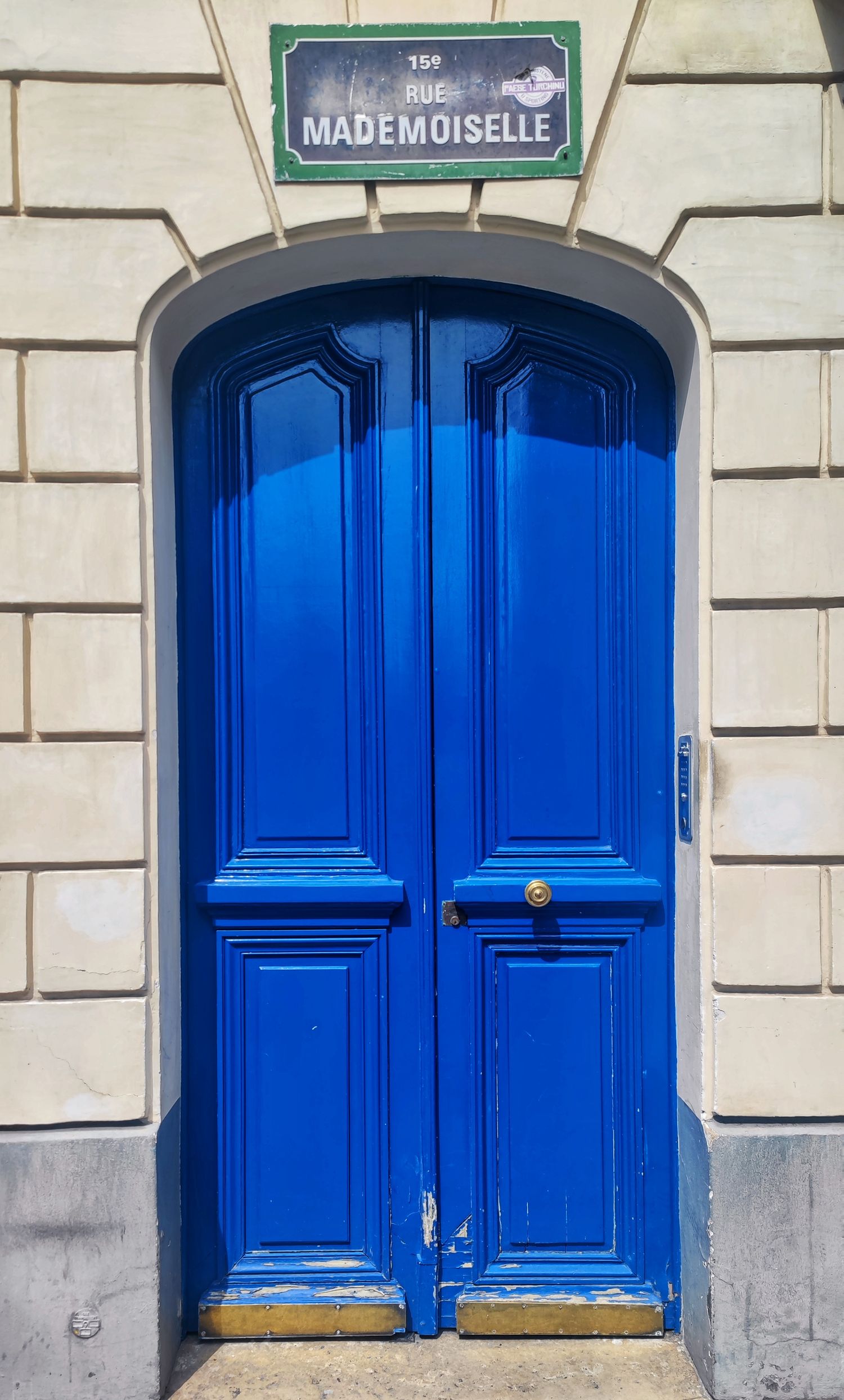 A bright blue arched double door in a cream stone wall beneath a green Rue Mademoiselle street sign in Paris.