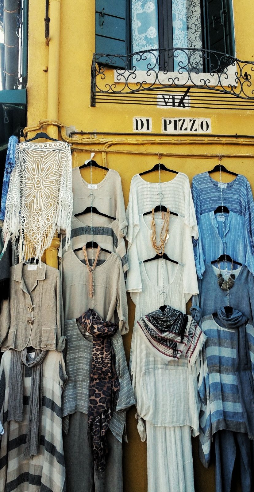 Linen blouses, dresses and scarves hang on a yellow wall below a wrought iron window and a Via di Pizzo sign.