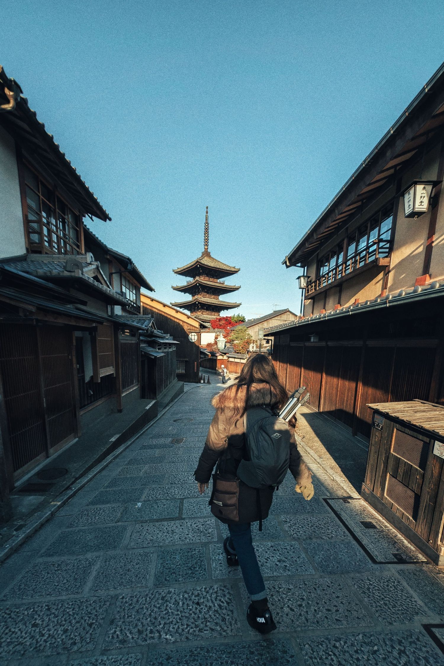 Woman with a backpack walks down a stone-paved street in Kyoto toward the Yasaka Pagoda, flanked by traditional wooden buildings.
