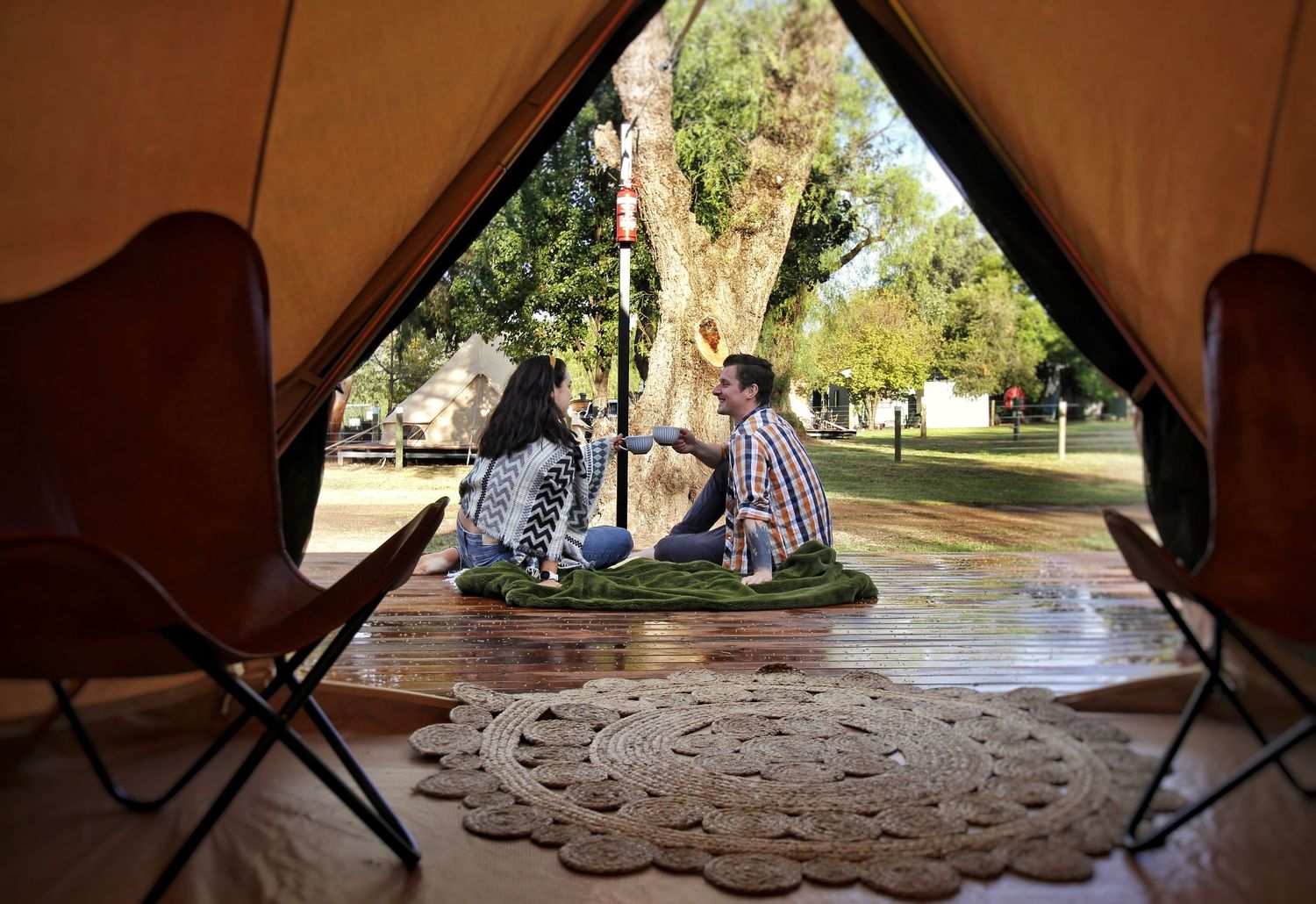 Two people sit on a round rug outside a canvas bell tent, viewed from inside past two leather butterfly chairs and a woven jute mat.
