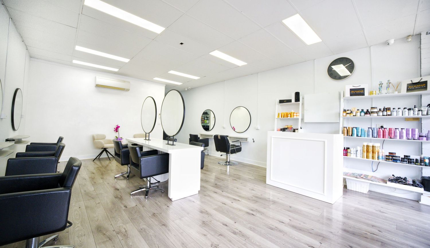 Spacious hair salon with black salon chairs, round mirrors, a white reception desk and shelves of hair care products on a white wall.